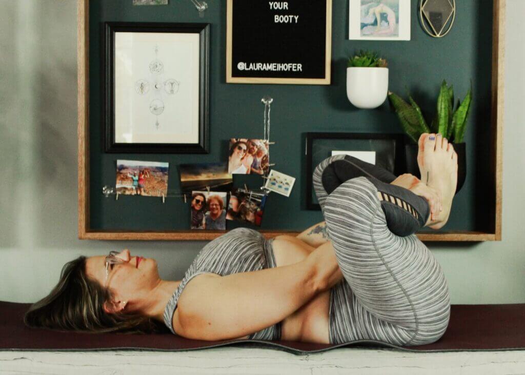 Laura on back hands holding both her ankles near pelvis knees bent out in gray yoga pants and top
