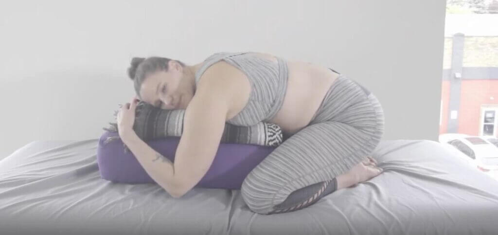 Laura on knees buttocks on feet chest rests forward on large purple yoga bolster and stacked blanket