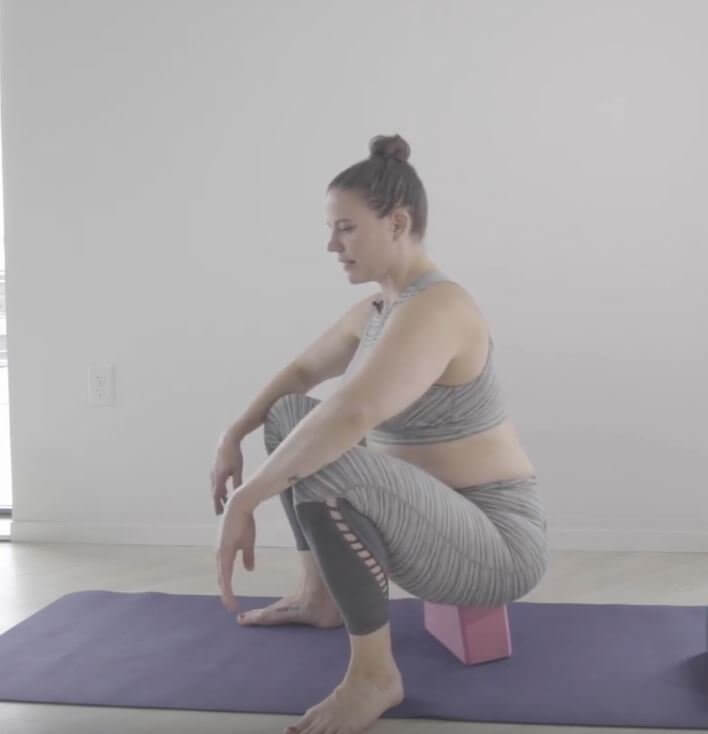 Feet apart she is in a squat knees bent tailbone resting on yoga block which is lain broad side down