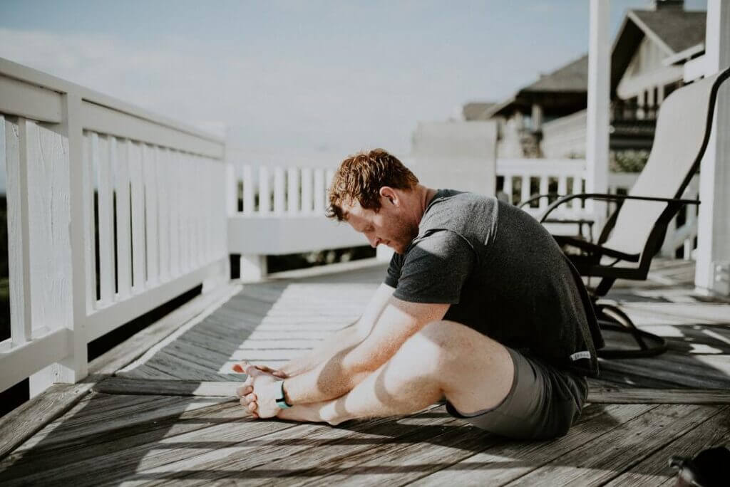 Man in workout clothes sitting on a wooden deck or outdoor terrace stretching his inner legs by pressing the soles of his feet together in a butterfly stretch