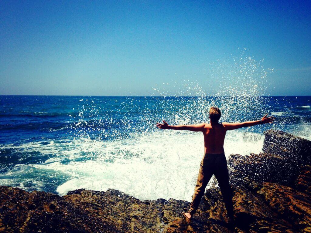 Man in pants no shirt barefoot standing on rocks facing ocean arms outstretched is splashed by wave