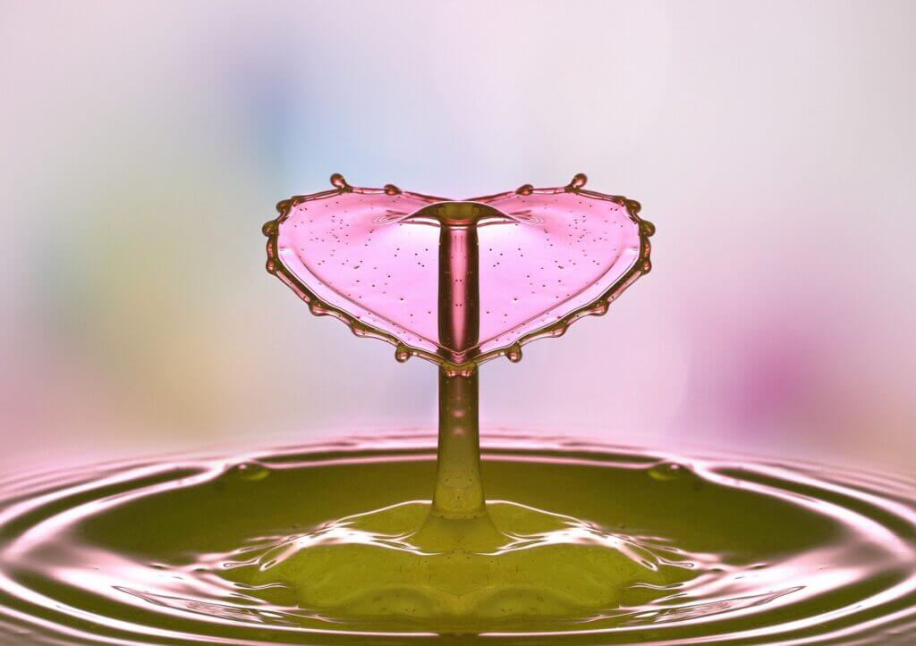 A droplet of water splashing in light pink water and the splash is in the shape of a pink heart