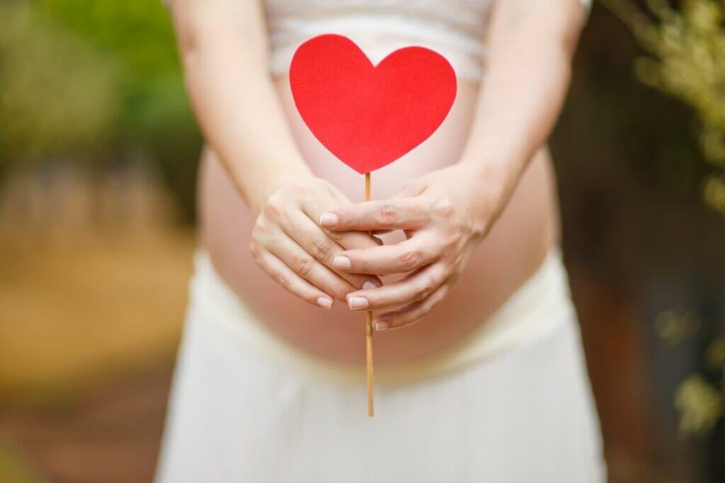 Blurry pregnant person in background holding red paper heart in foreground in front of exposed belly