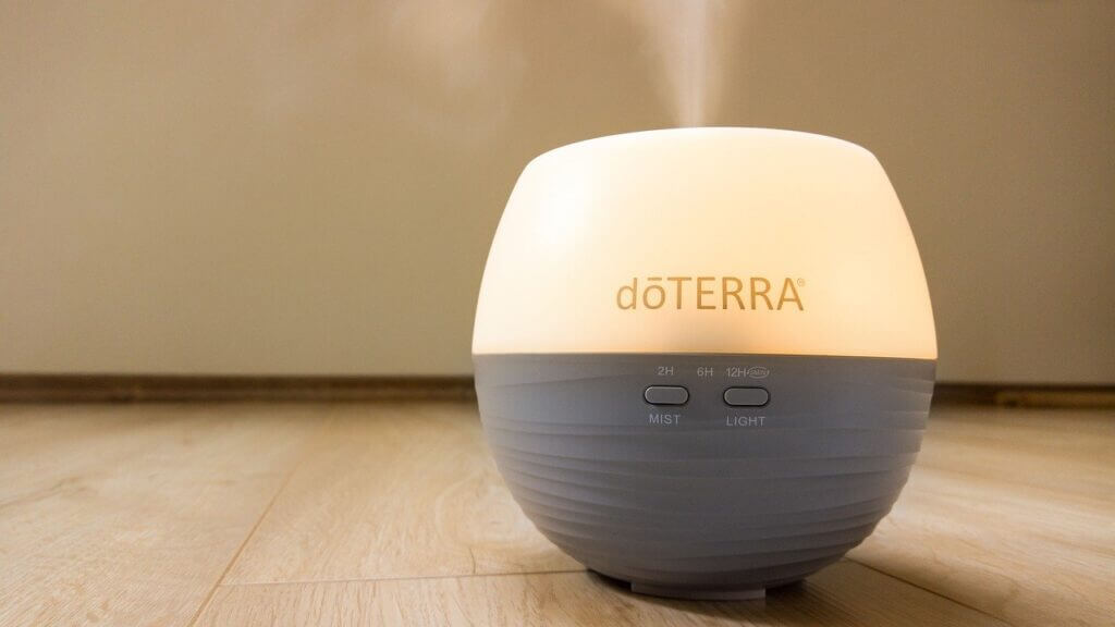 Grey DoTerra diffuser sitting on light colored wooden floor turned on with steam coming out the top