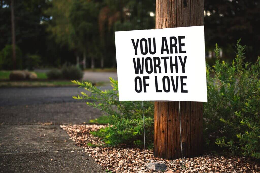 White yard picket sign in front of a large wooden telephone pole reads you are worthy of love