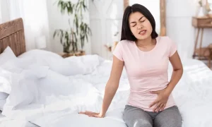 Woman sitting on a bed holding her pelvis with a pained expression on her face